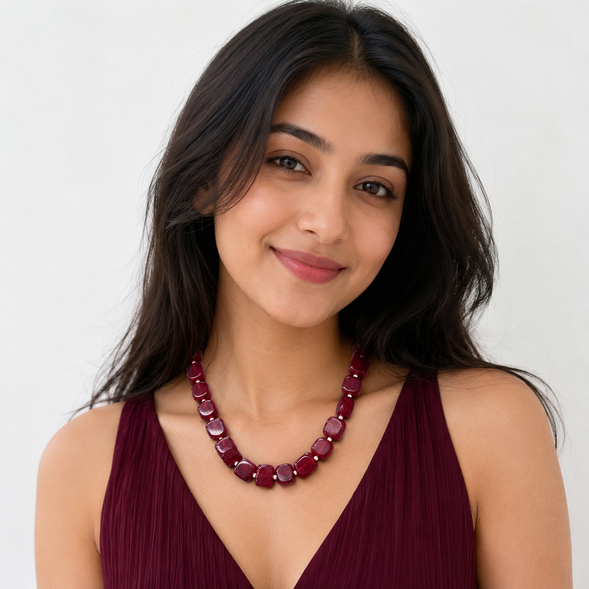 A woman with long dark hair wearing a chunky maroon beaded necklace and a matching maroon top, photographed against a clean white background.