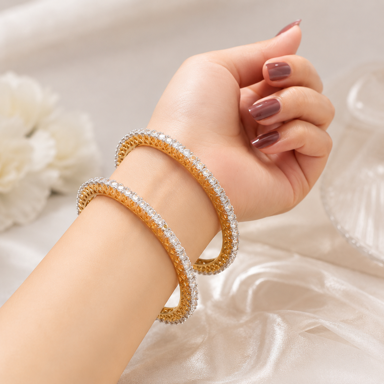 Two gold-toned bangle bracelets, one paved with clear crystals and the other with clear crystals and white stones, worn on a woman's wrist against a soft white satin background.