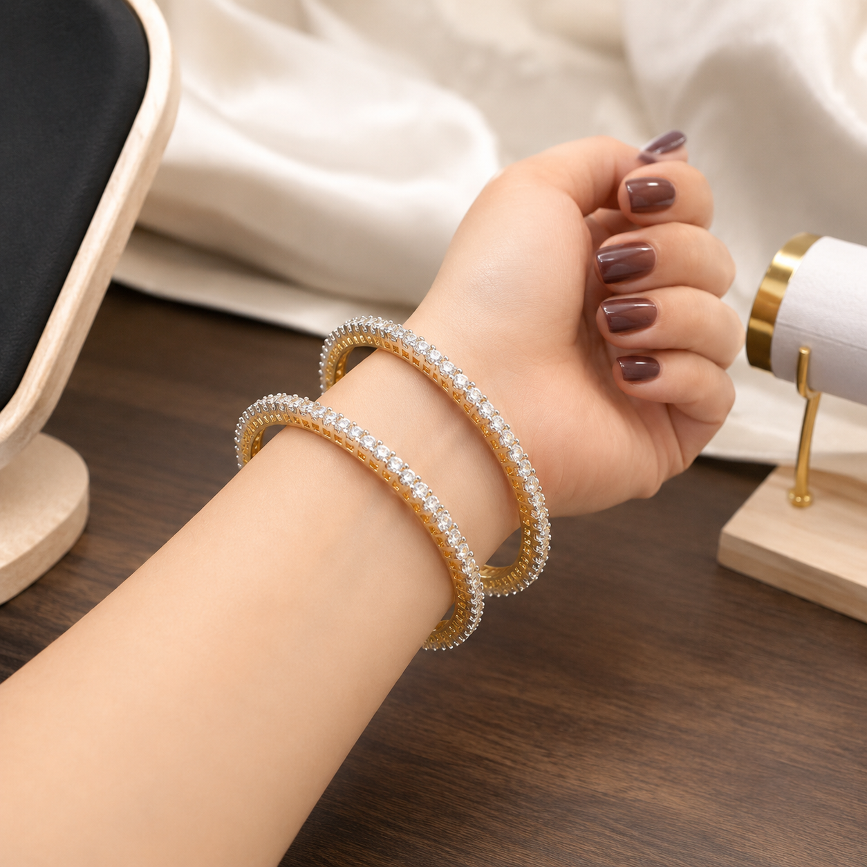 A close-up of a person's wrist wearing two gold bangles adorned with sparkling clear stones, set against a soft cream fabric and dark wooden tabletop.