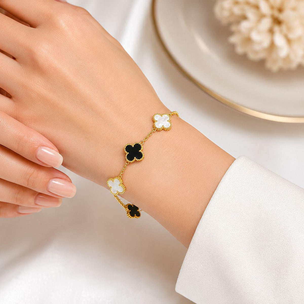A gold-tone bracelet with alternating white and black clover motifs worn on a woman's wrist, shown against a soft white background with a plate in the blurred background.
