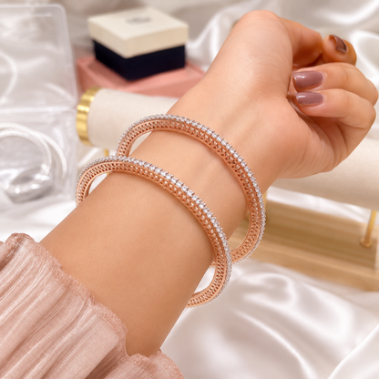 A close-up of a person's wrist wearing two thin braided bracelets, one rose gold and one silver, both set with clear stones. The background features soft white satin fabric and blurred jewelry boxes.