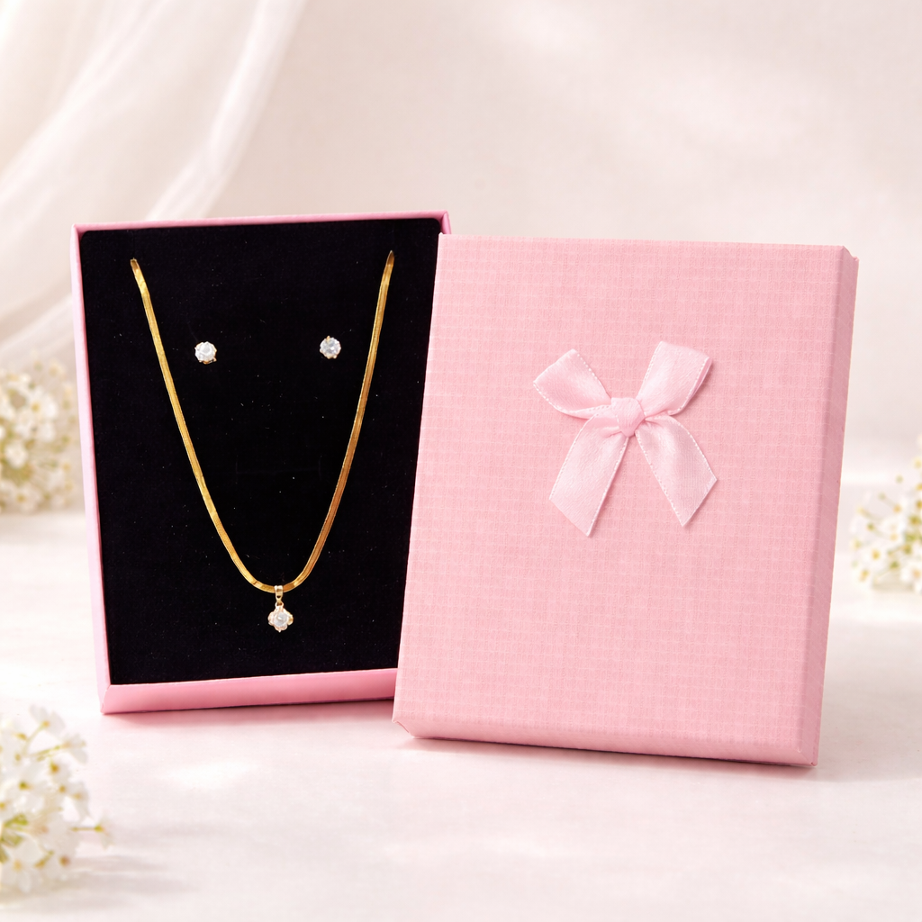 Gold necklace and earrings on a black card next to a pink gift box with a bow.
