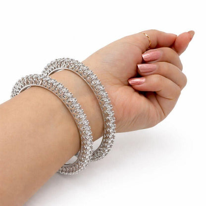 A close-up of a person's wrist wearing three thick silver-toned bangles covered in sparkling clear stones, with a slim gold ring on the finger, against a plain white background.