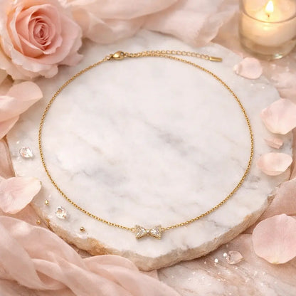 Gold necklace with a bow pendant on a marble surface surrounded by pink roses and petals.