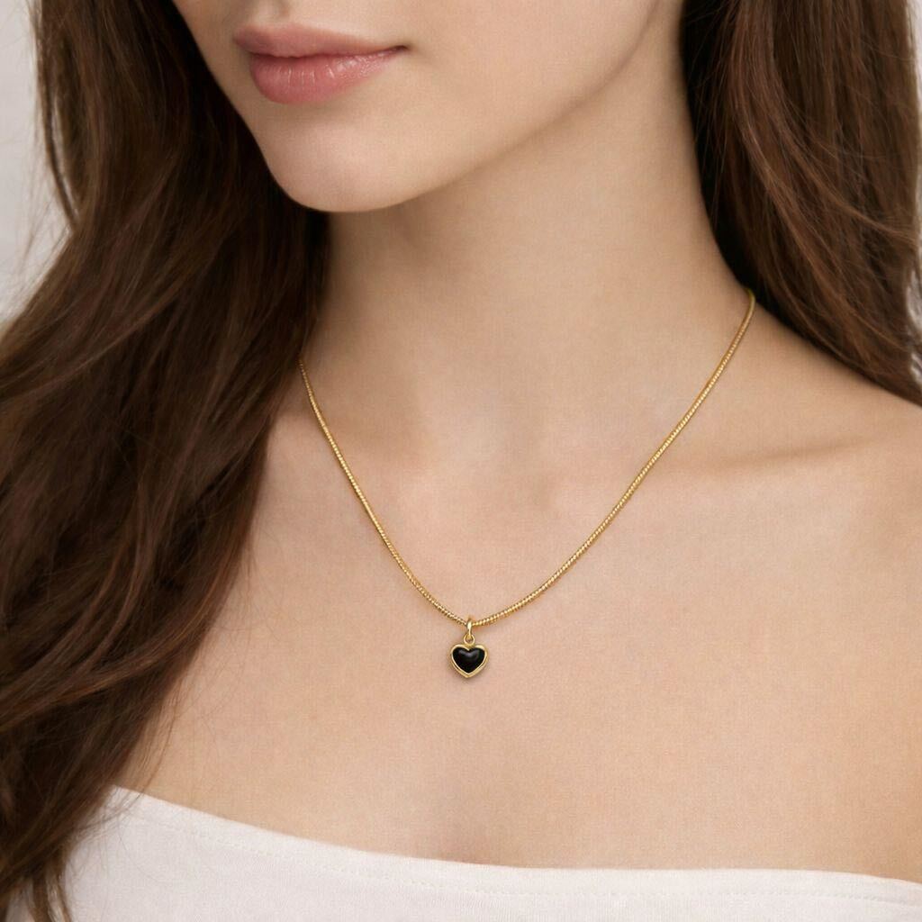 A close-up of a woman wearing a delicate gold chain necklace with a small heart-shaped pendant featuring a dark green inlay.