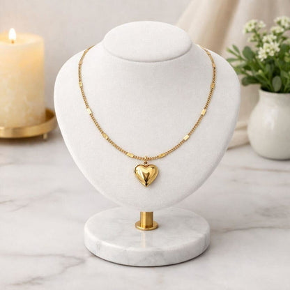 Gold necklace with a heart pendant on a marble stand, candle and plant in the background